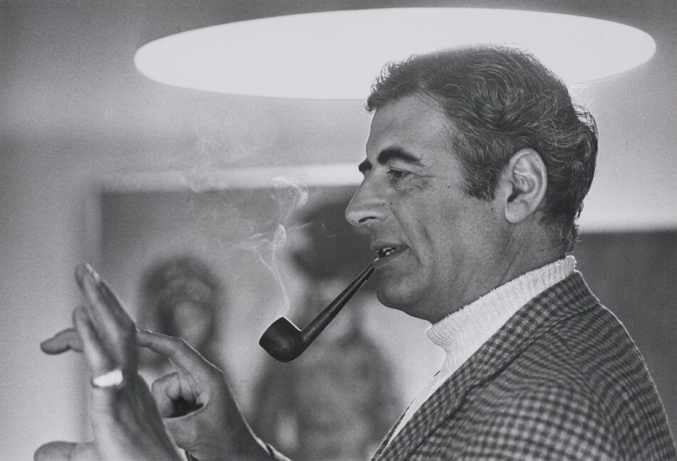 A black-and-white photo of jewelry designer Andrew Grima smoking a pipe in his home studio in 1969