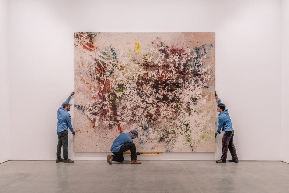 Three men in gloves and blue workshirts hang a large-scale square artwork on a white wall at the downtown Los Angeles location of art shipping and storage company UOVO.