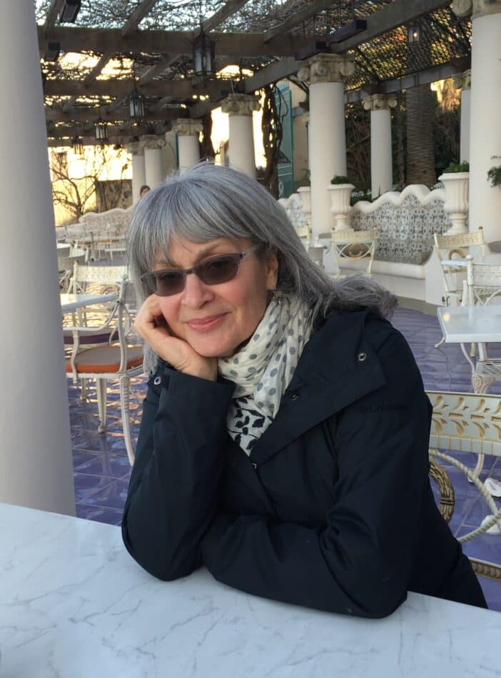 Author Cara Greenberg wearing a jacket, scarf and sunglasses and sitting at a table outdoors