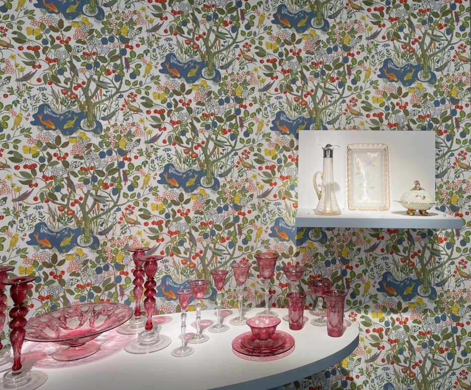 A display in the exhibition "Brilliant Color" at the Corning Museum of Glass showing colorful glassware against a wallpaper design with cherry trees, goldfish, birds and yellow butterflies against a white background