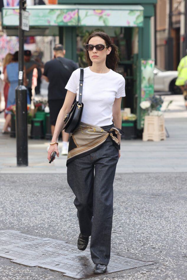 Alexa Chung on a London street wearing dark sunglasses, a white T-shirt and dark jeans with a scarf tied around her waist.