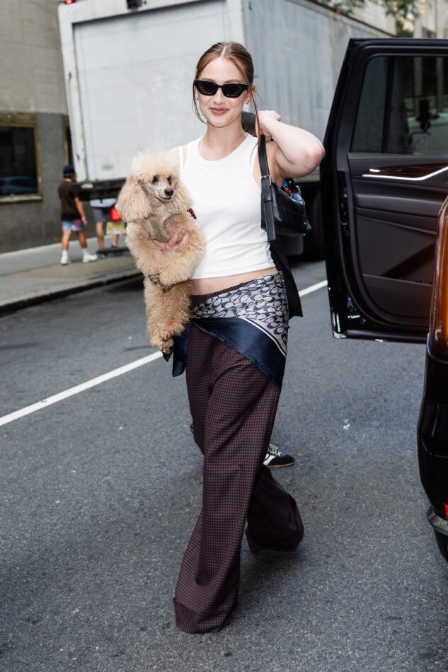 Lola Tung on a New York street carrying a dog and wearing dark sunglasses, a white tank top, dark checkered trousers and a scarf tied around her waist