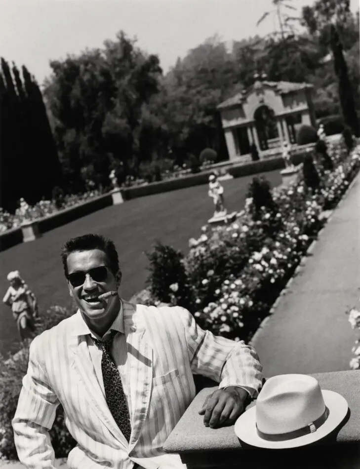 Black-and-white photo of Arnold Schwarzenegger sitting in a garden smoking a cigar