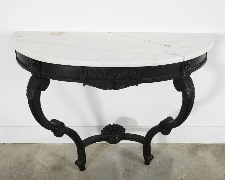 An ornate Louis XV–style demi-lune console table with a white marble top and ornate black legs stands against a blank white wall. 
