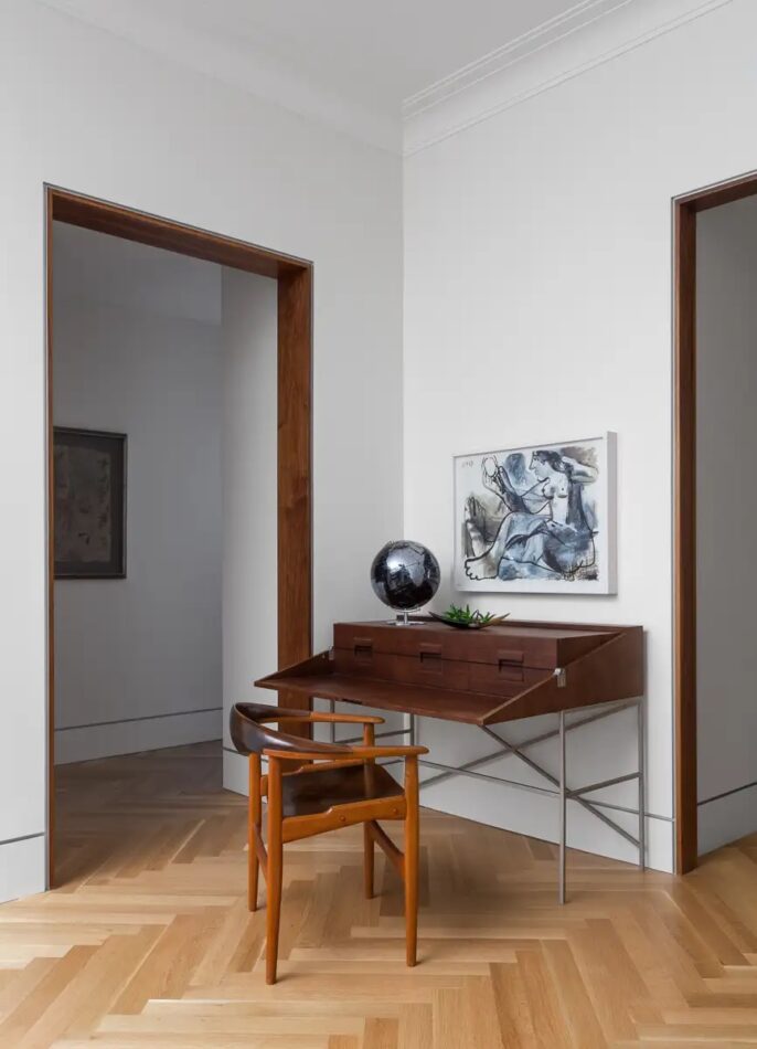 A white-walled office with a small wooden secretary on metal legs and a modern artwork depicting a nude figure in blues and grays hanging just above it