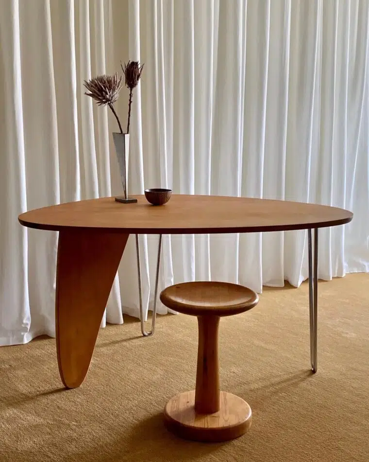 An Isamu Noguchi for Herman Miller Rudder table paired with a wooden stool in front of a wall of white curtains