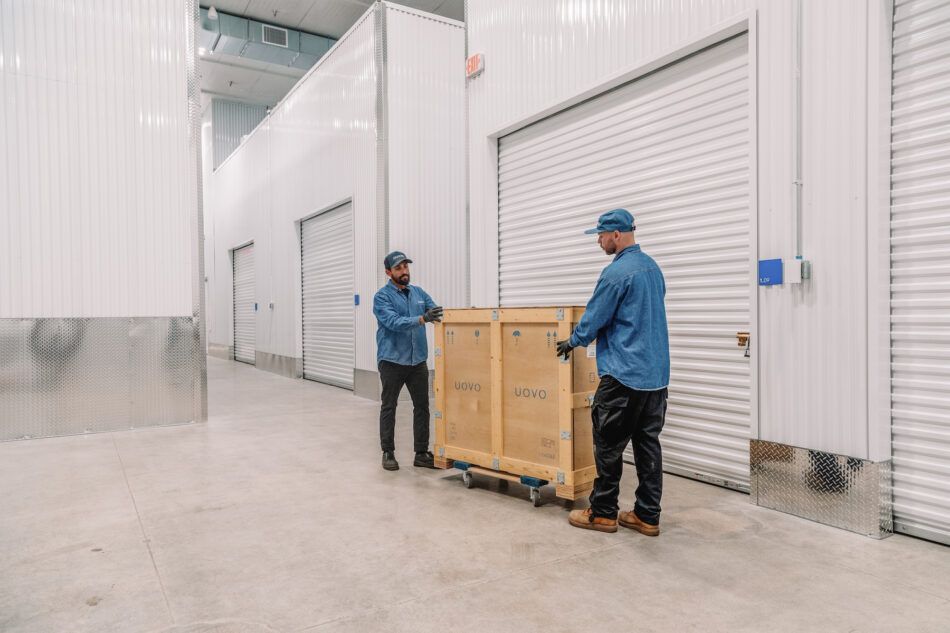 Two men in gloves and blue workshirts wheel a wooden crate down a hallway of storage units with corrugated white walls at the downtown Los Angeles location of art shipping and storage company UOVO.