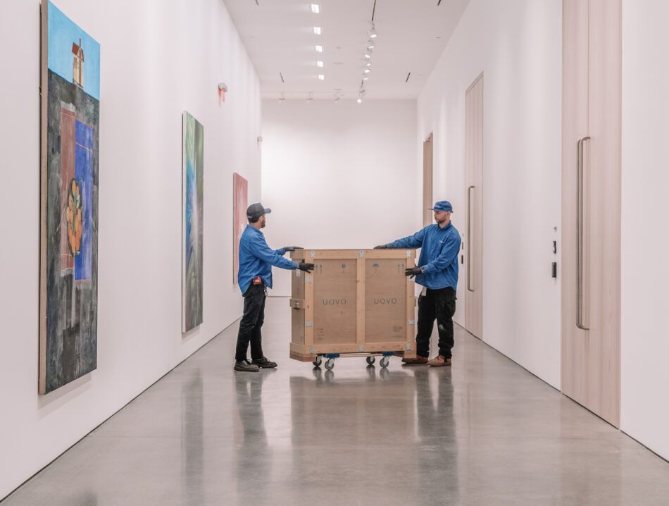 Two men in gloves and blue workshirts wheel a wooden crate down a hallway hung with art at the downtown Los Angeles location of art shipping and storage company UOVO.