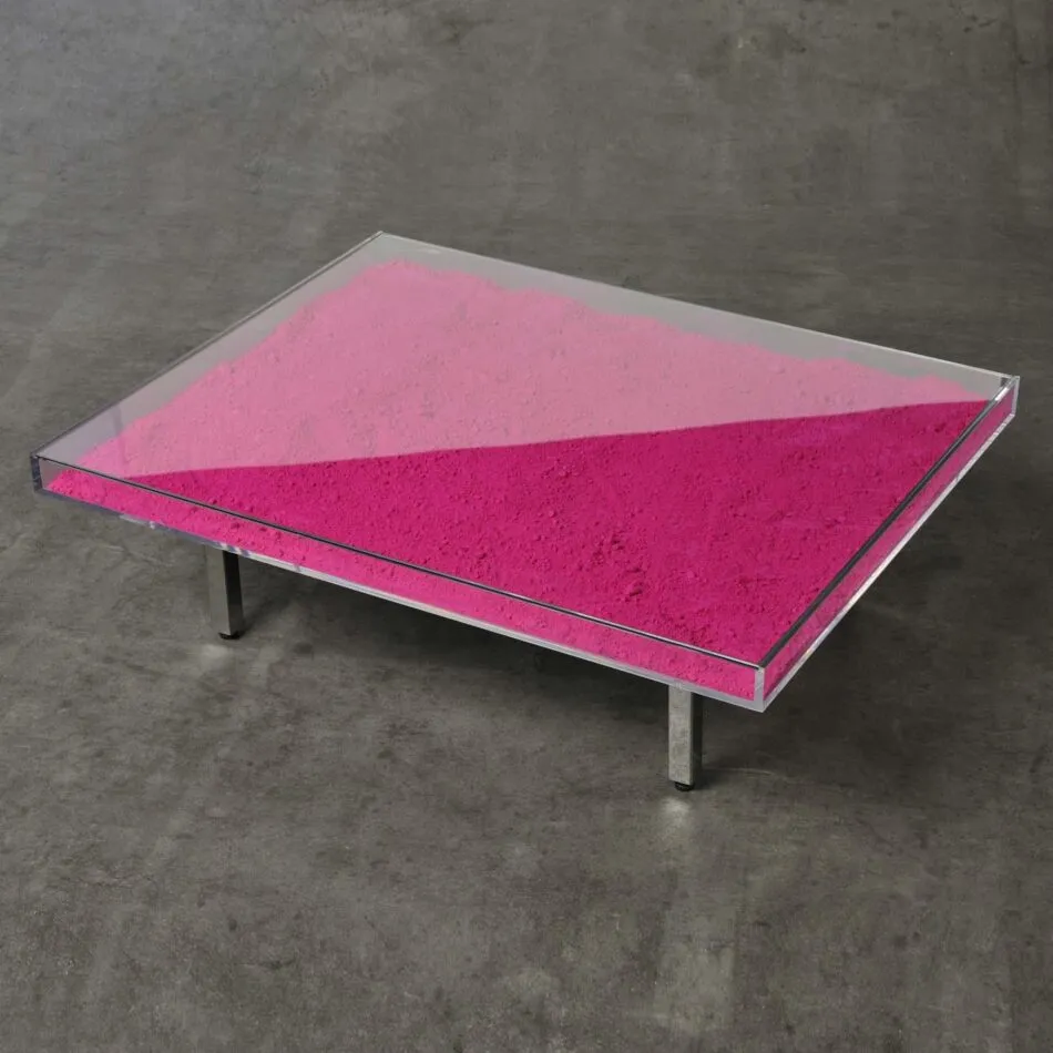 An Yves Klein coffee table with metal legs and loose pink pigment powder inside a clear square tabletop sits on a concrete floor. 