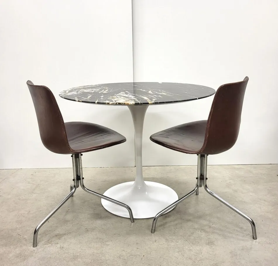 An Eero Saarinen Tulip table with a white pedestal base and marbled-black top, flanked on both sides by brown chairs with metal legs.