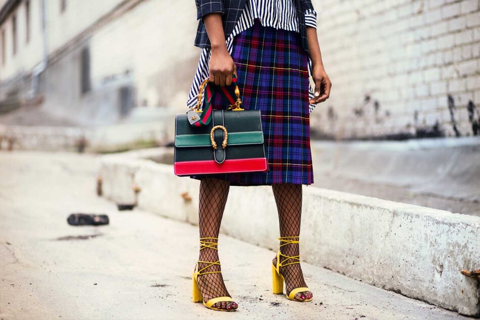 fashionable woman in a plaid skirt from the waist down holding a red and green bag with yellow heels on.