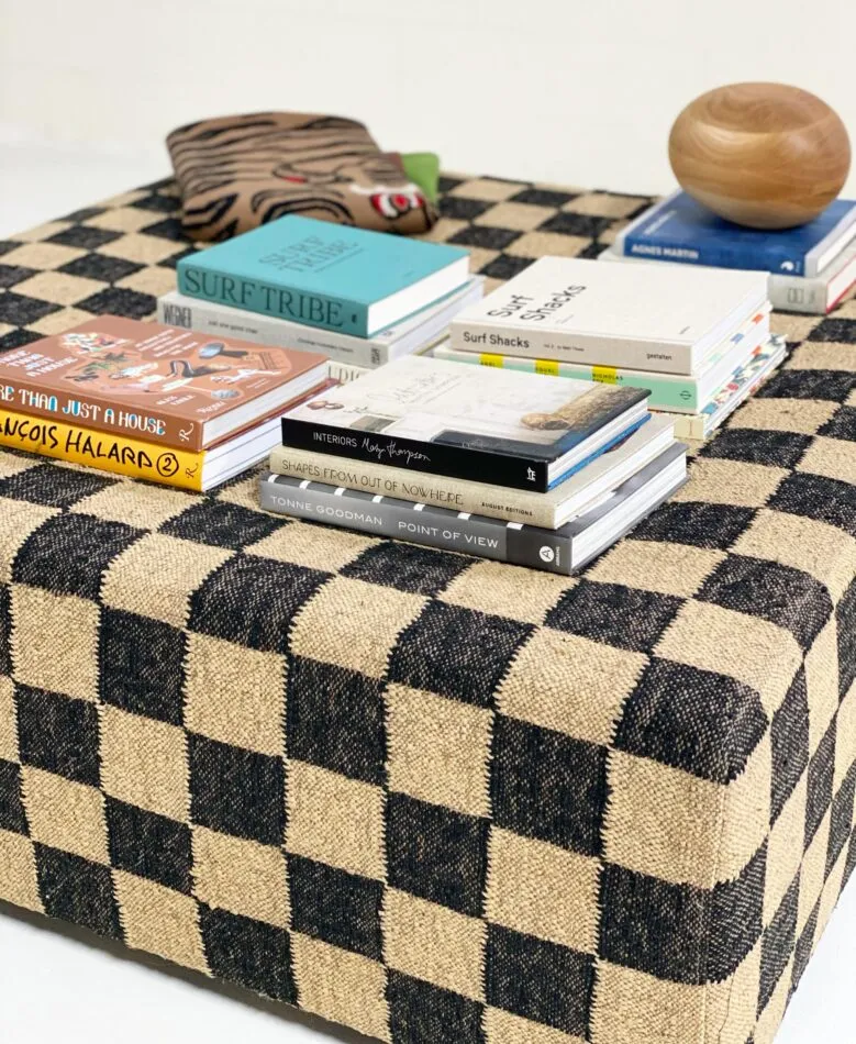 A selection of brightly-colored coffee table books sit atop a square ottoman covered in tan and black checkered upholstery. 