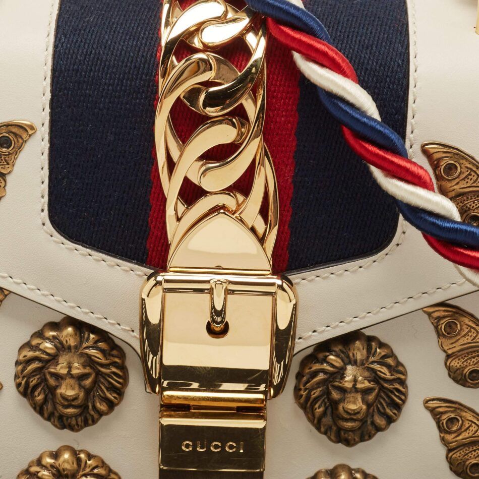 A close-up of the gold metal latch on a beige Gucci Sylvie bag, with gold lion and butterfly studs flanking it on either side and a red, white and blue braided strap draped over the flap. 
