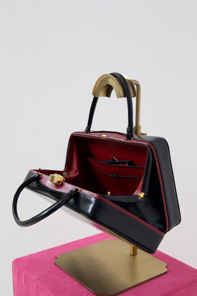 An open black-leather 1950s Gucci handbag hangs by one of its handles on a gold stand, showcasing its bright red lining. 