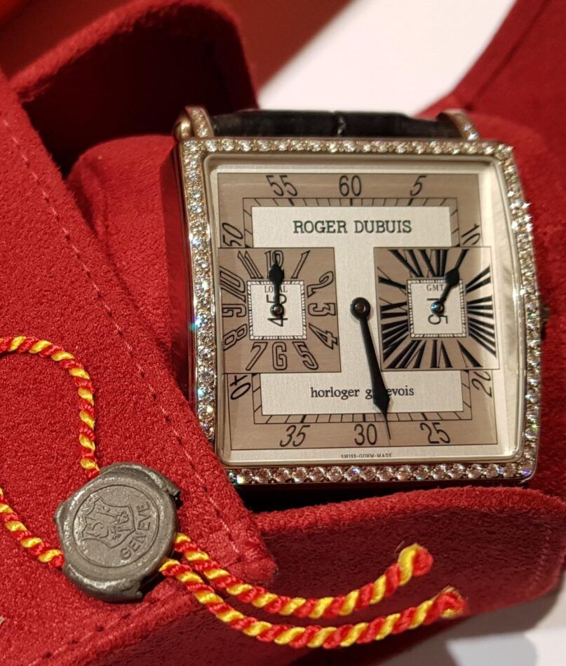 A Roger Dubuis watch featuring a black alligator strap and a square face that has both Arabic and Roman numerals and is surrounded by diamonds, presented in it's original red box 